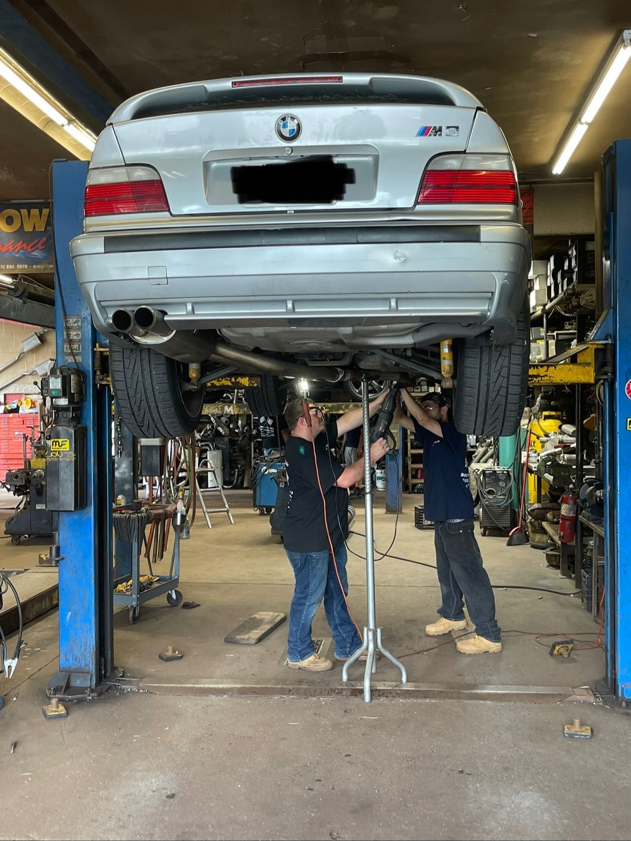 Two mechanics working under a silver car on a lift in an auto repair shop