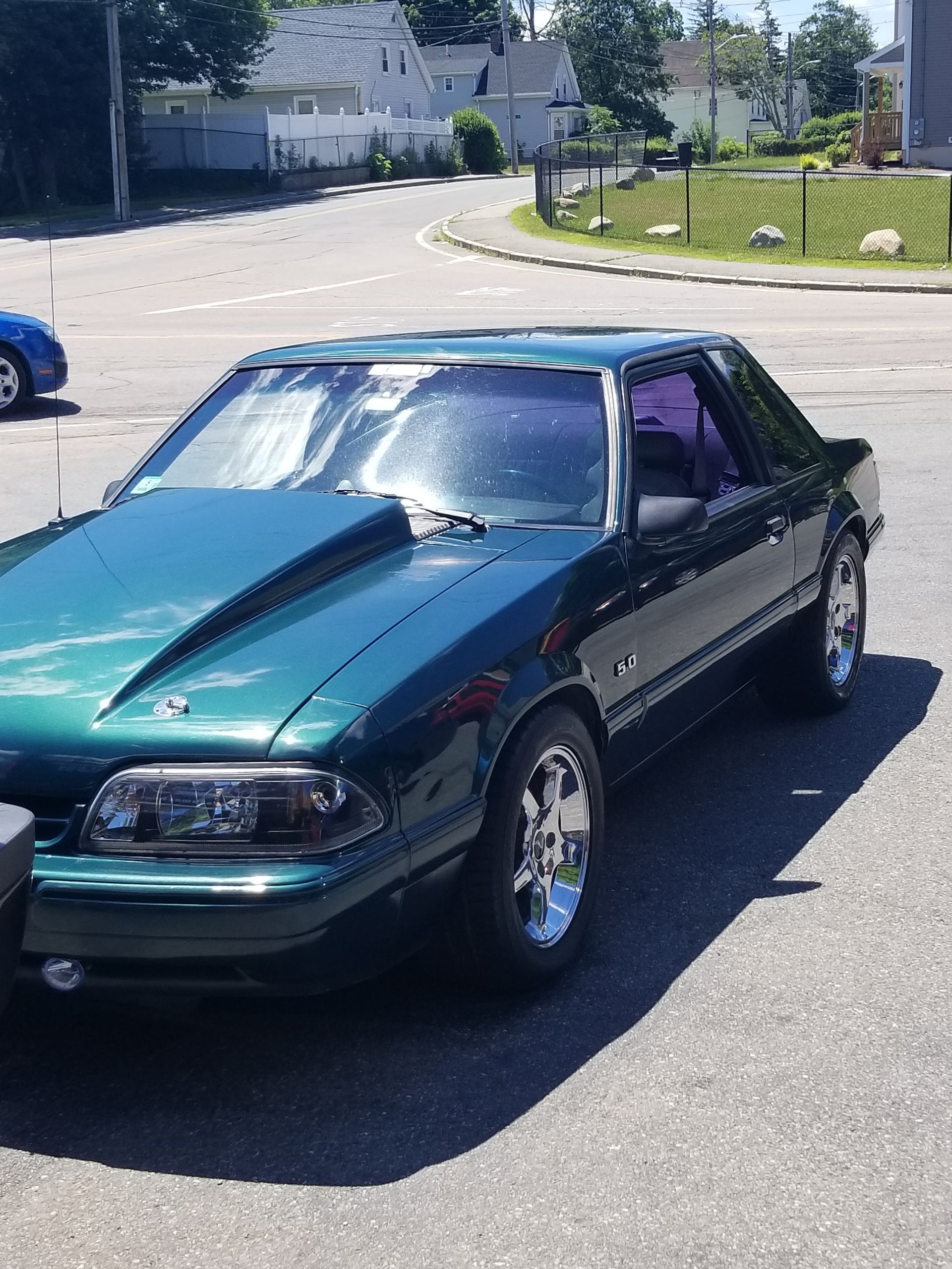 Teal sports car parked on a sunny street near a grassy curb and houses