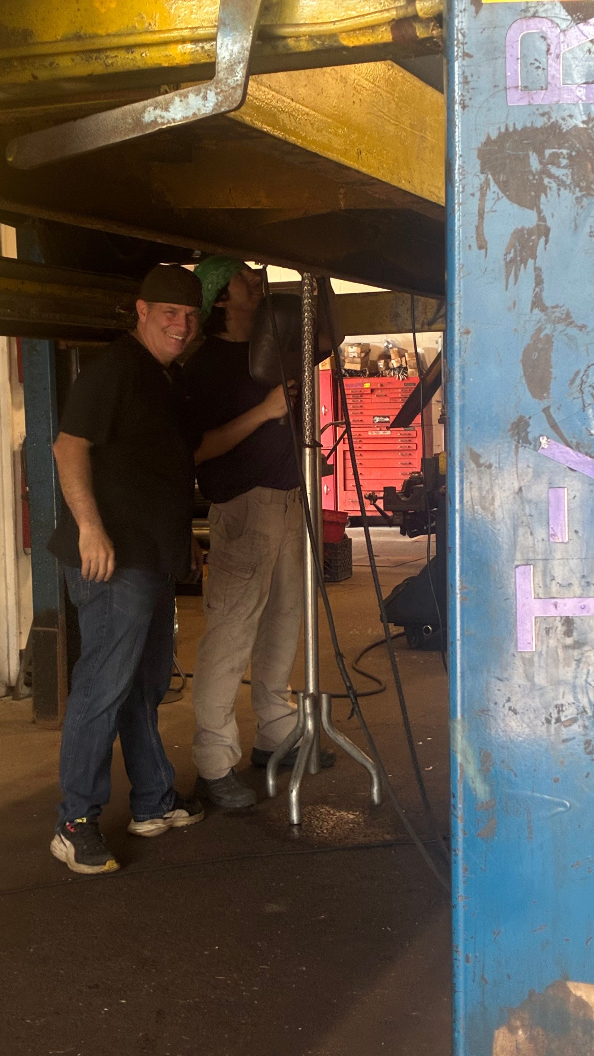 Two people standing under a raised vehicle, one holding a jack in a workshop.