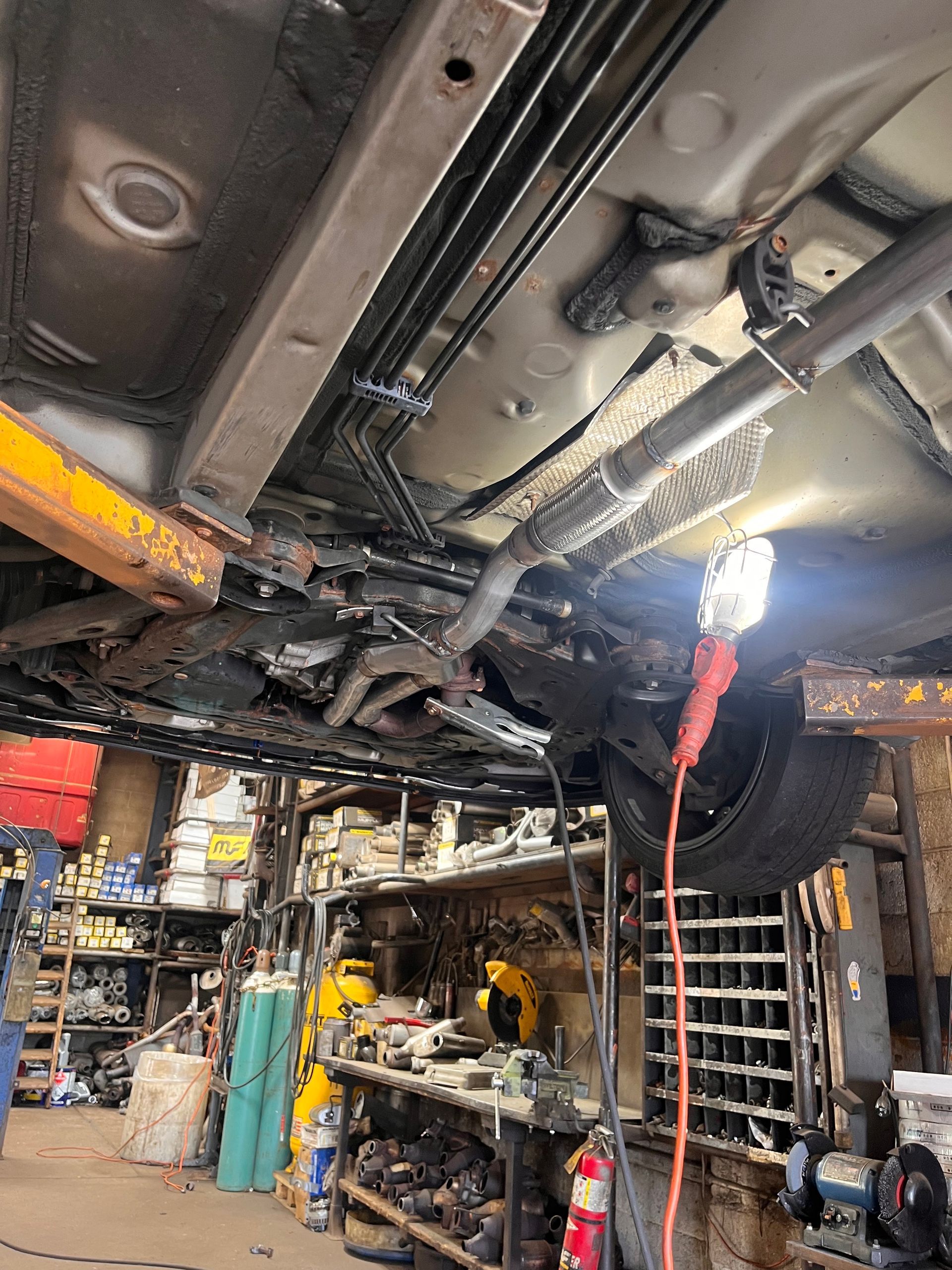 Car underside on a lift in an auto repair shop, with tools, shelves, and a mechanic’s work light nearby.