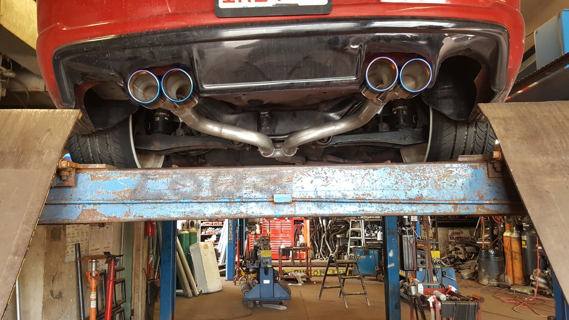 Rear view of a red car on a lift in an auto repair shop, with four chrome exhaust tips visible.