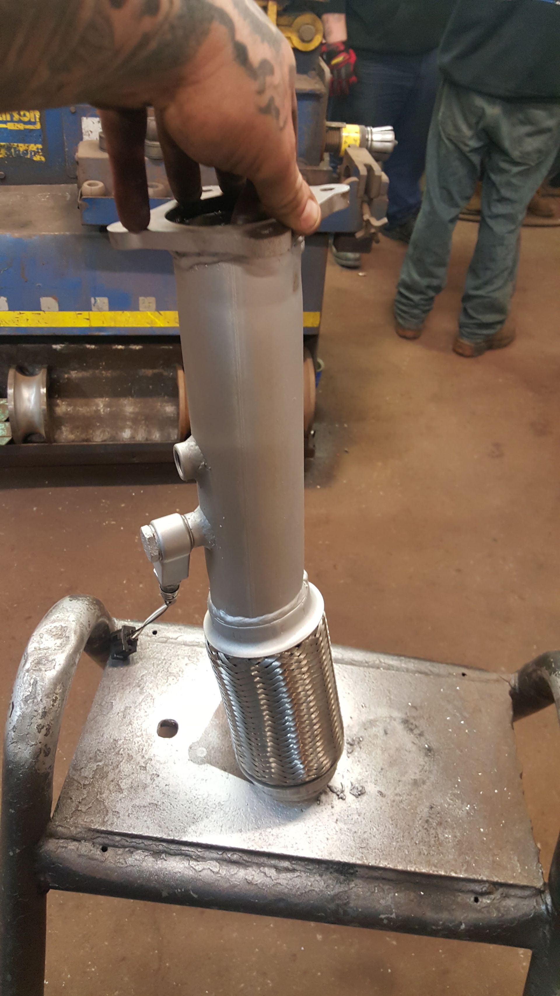 Hand holding a gray pipe on a metal workbench with a shiny perforated fitting and tools in the background