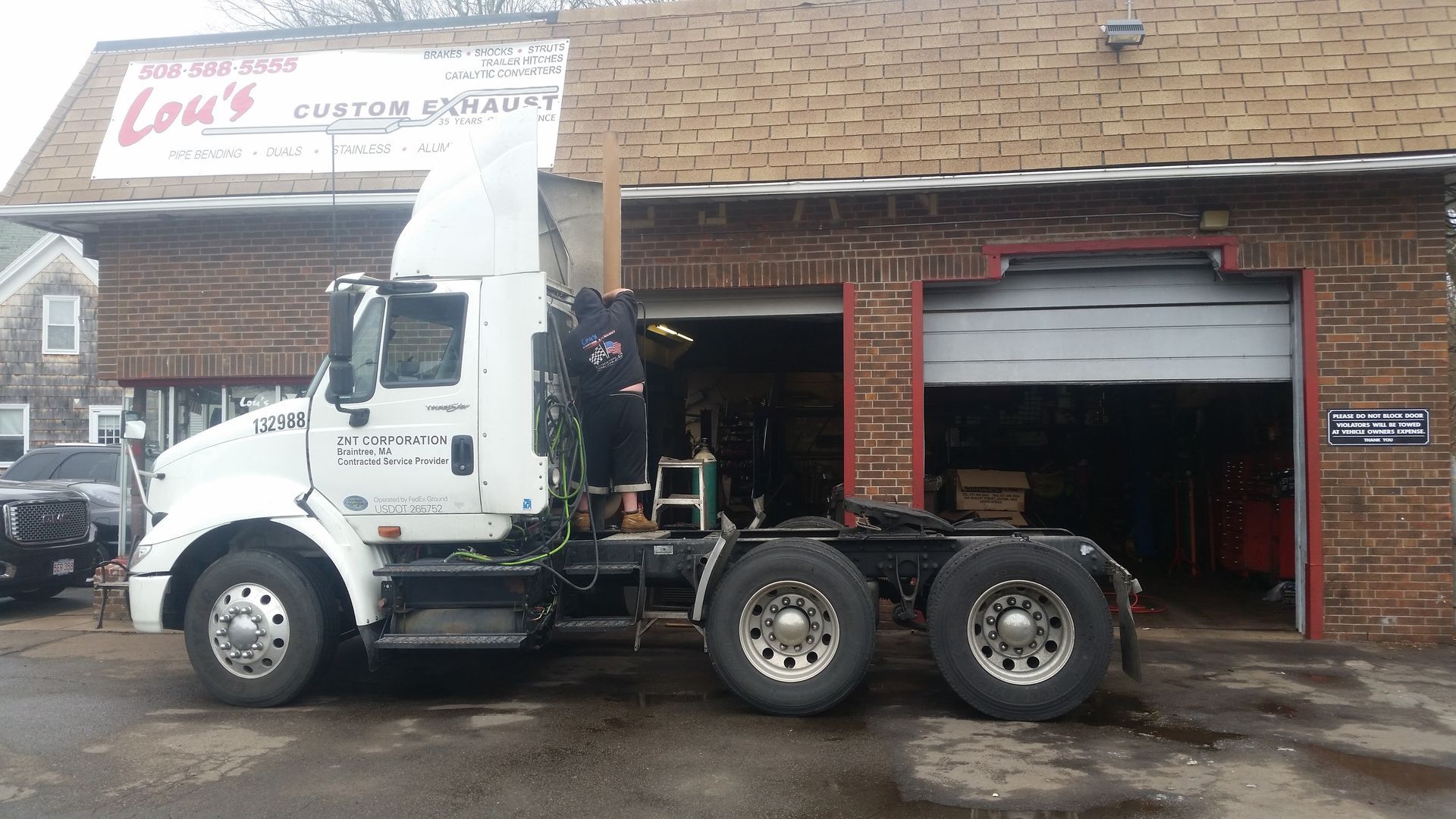 White semi-truck parked outside a brick auto shop with open bay doors on a wet street