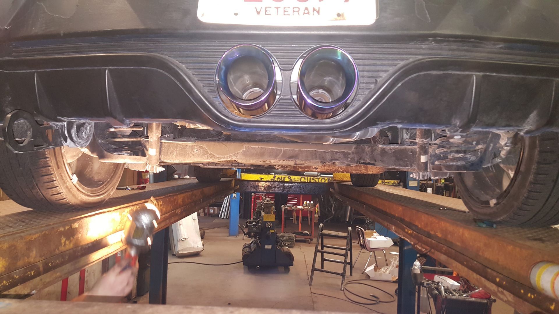Underbody view of a black car on a lift, showing dual exhaust tips in an auto repair shop