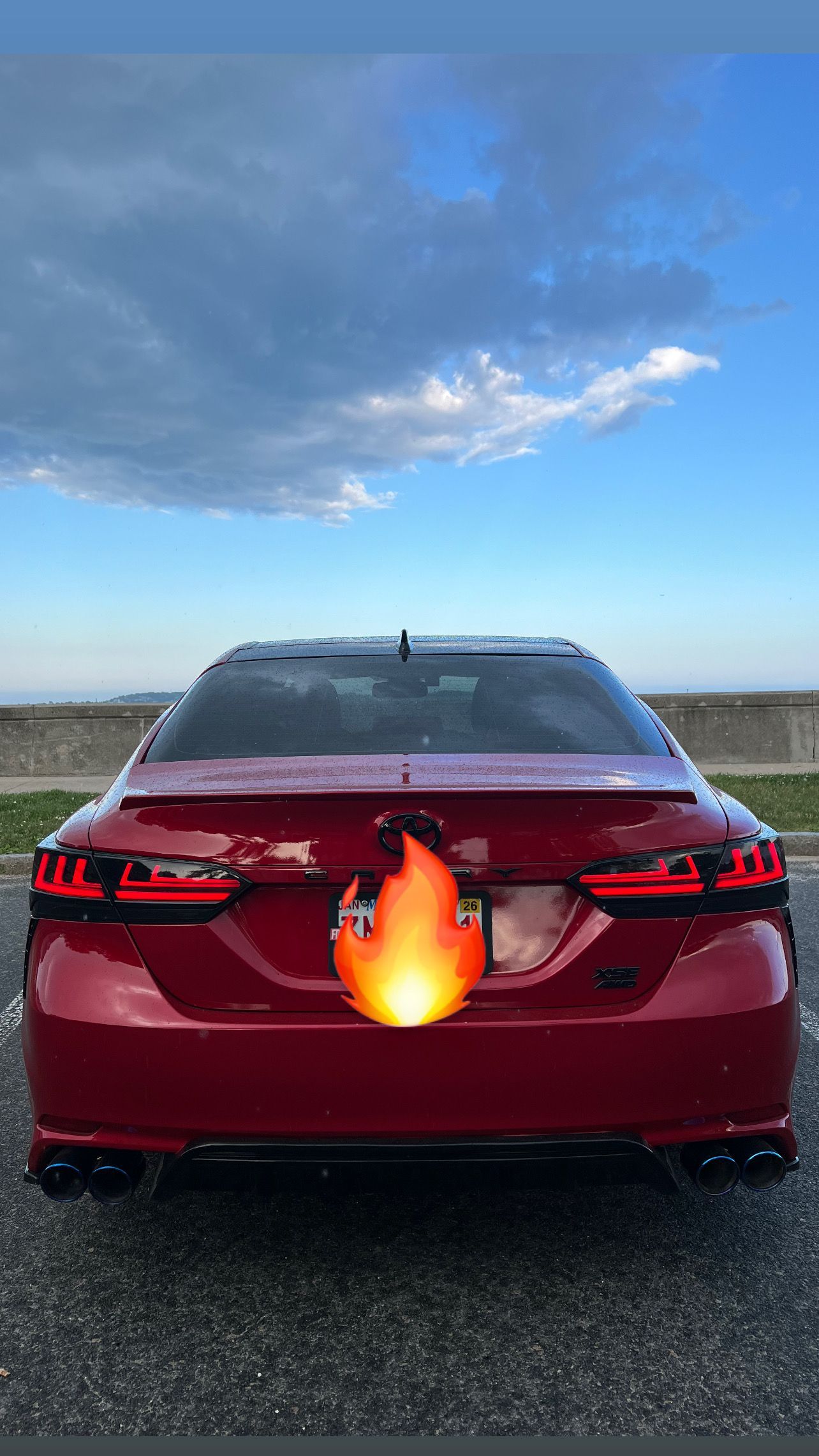 Rear view of a red sedan on a road under a cloudy sky, with a flame sticker over the license plate area