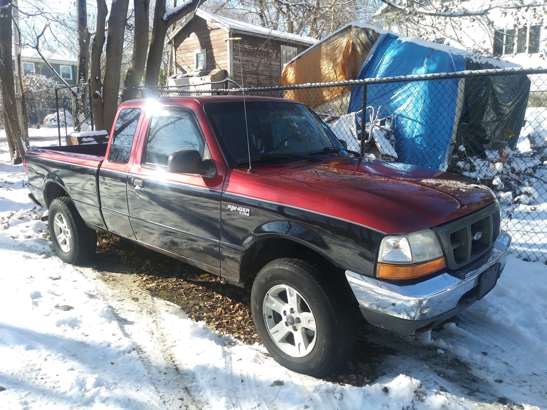 Red pickup truck parked in a snowy driveway beside a blue tarp-covered structure