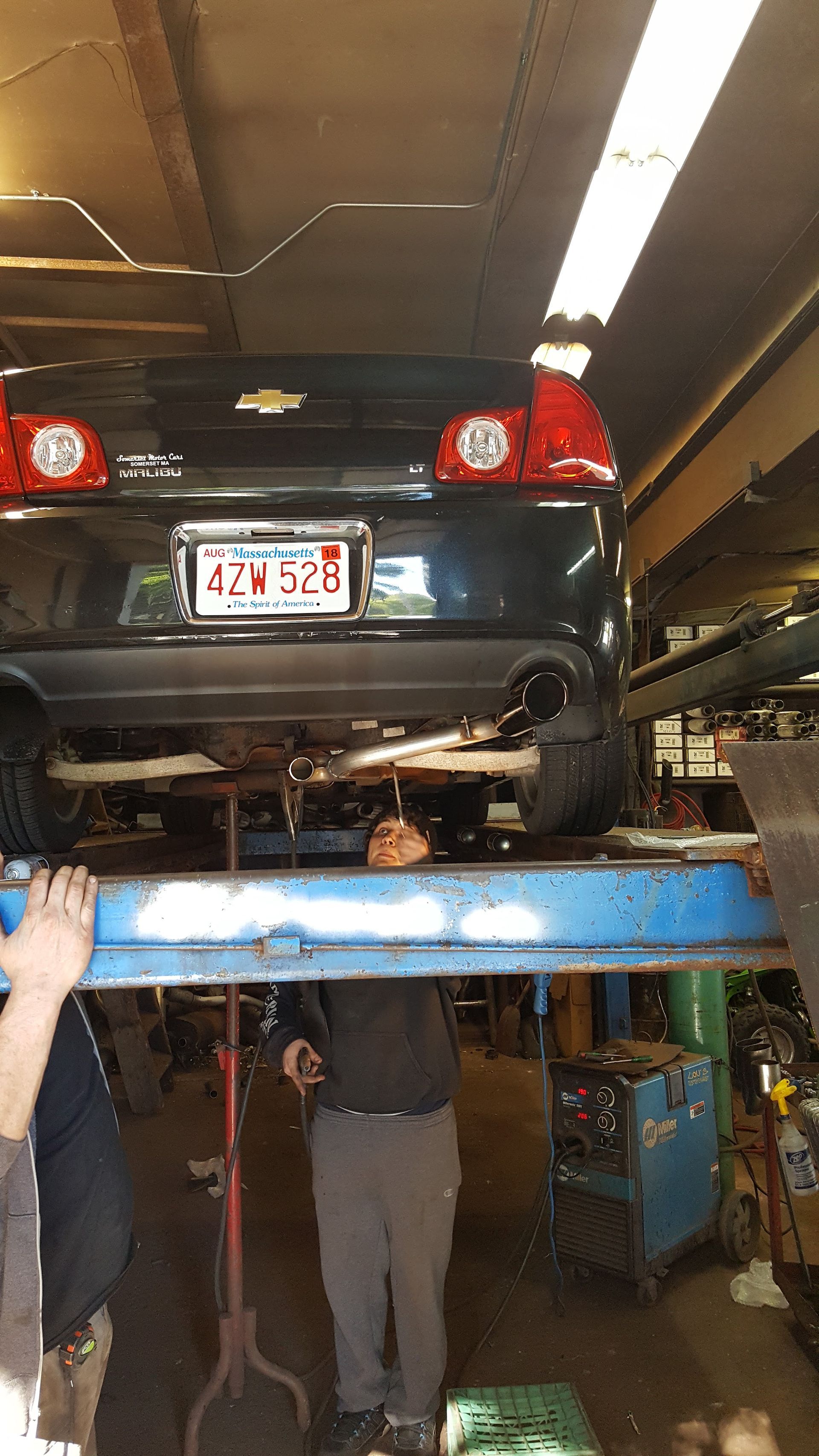 Car lifted on a blue auto hoist in a garage, with two mechanics underneath and a hand at left.