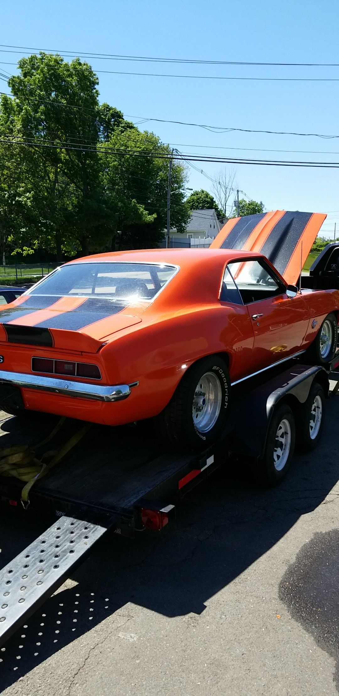 Orange classic car on a tow trailer, rear view, with ramps extended on a sunny road.