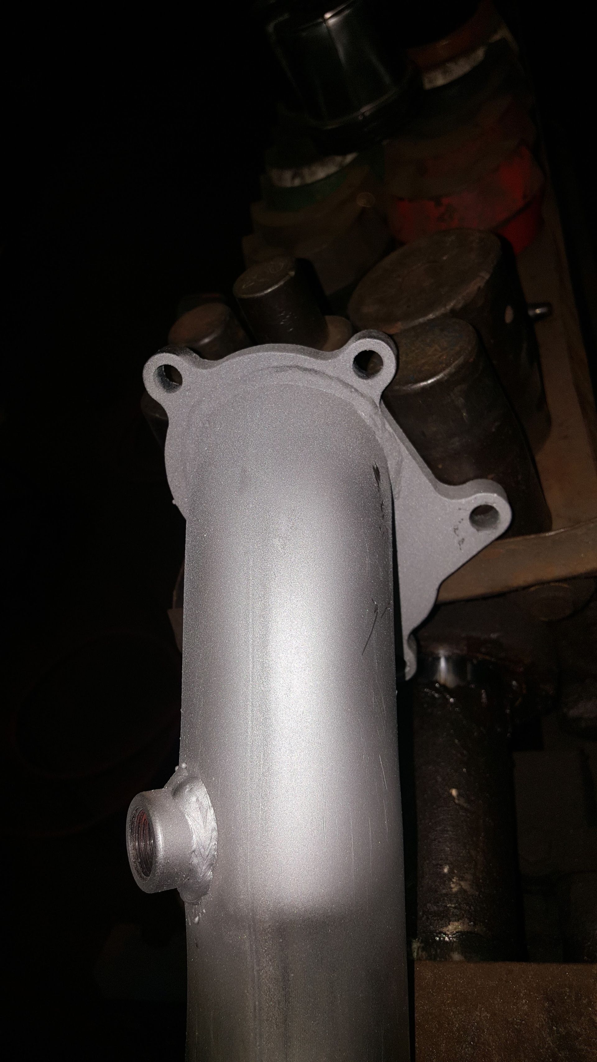 Silver metal exhaust manifold with mounting holes in a dark workshop setting