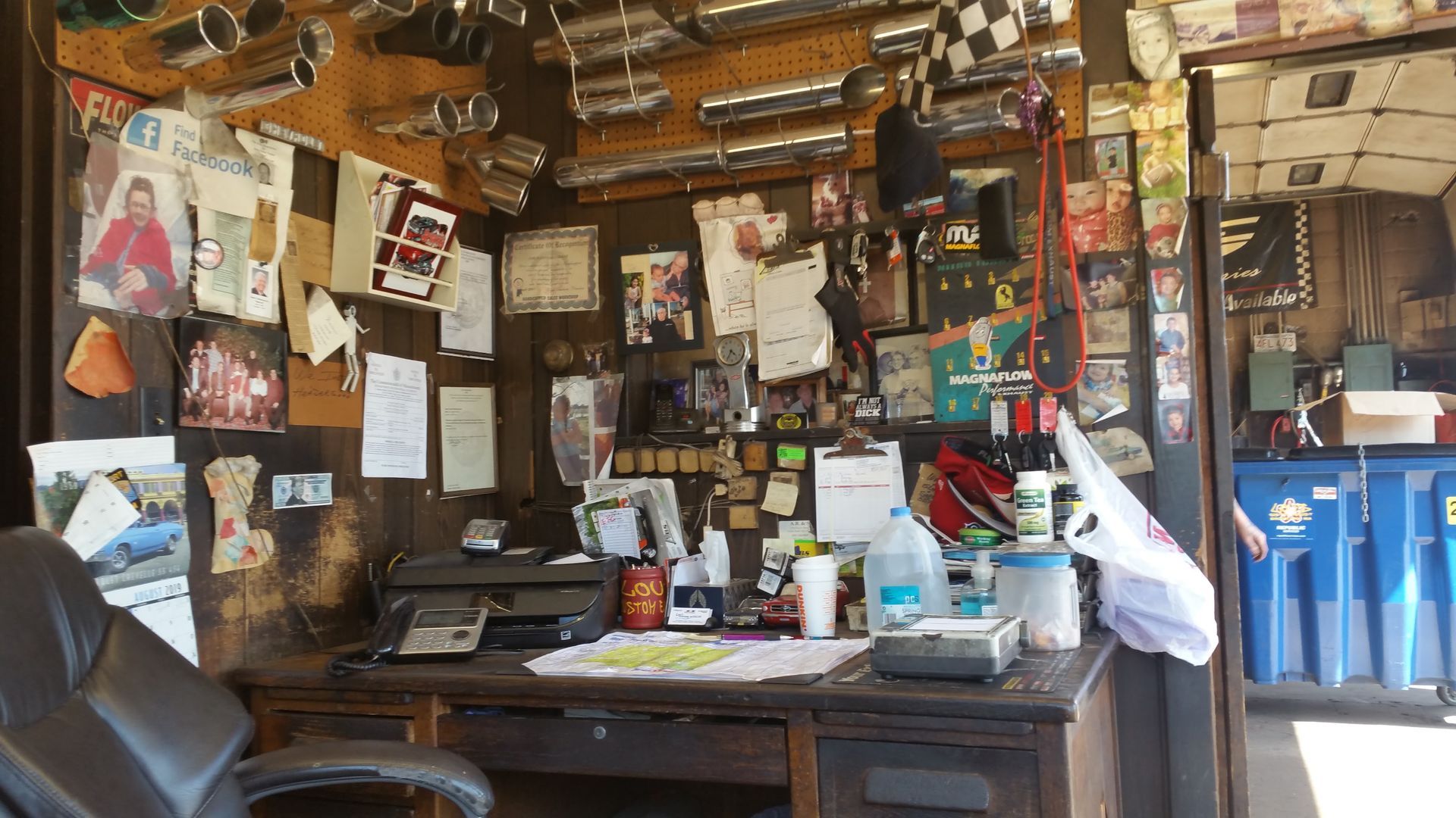 Cluttered workshop desk with tools, papers, and hanging items under a wooden ceiling.
