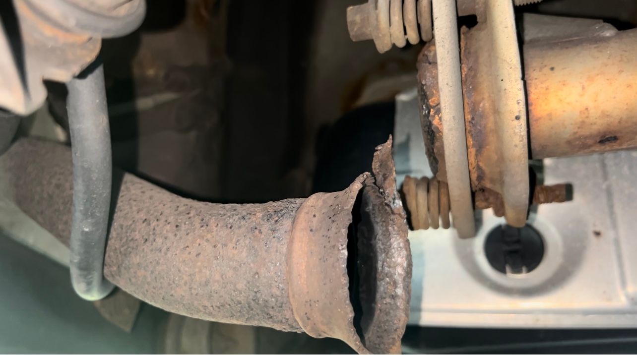 Rusty exhaust pipe with a broken, leaking connection near a vehicle undercarriage