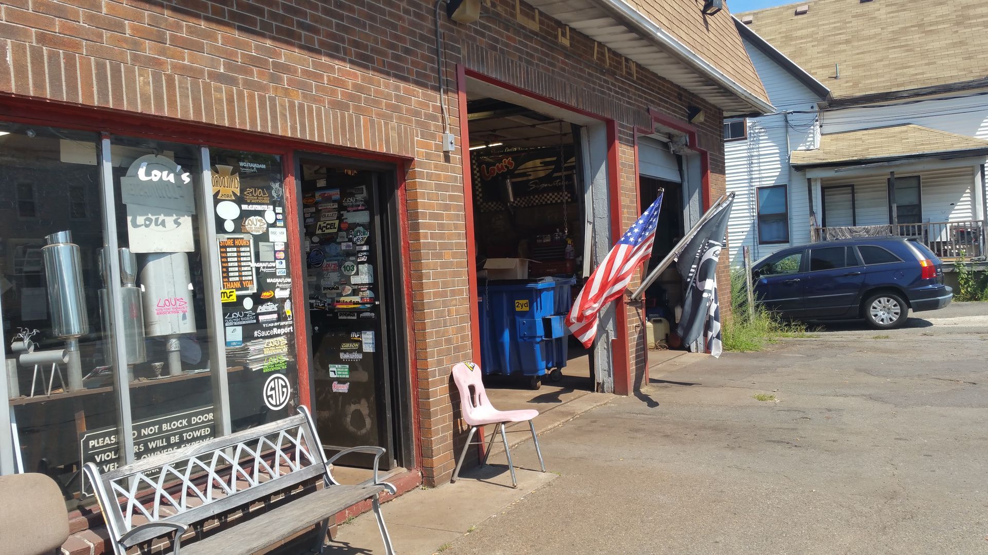 Small brick storefront with a flag display and parked car outside on a sunny day