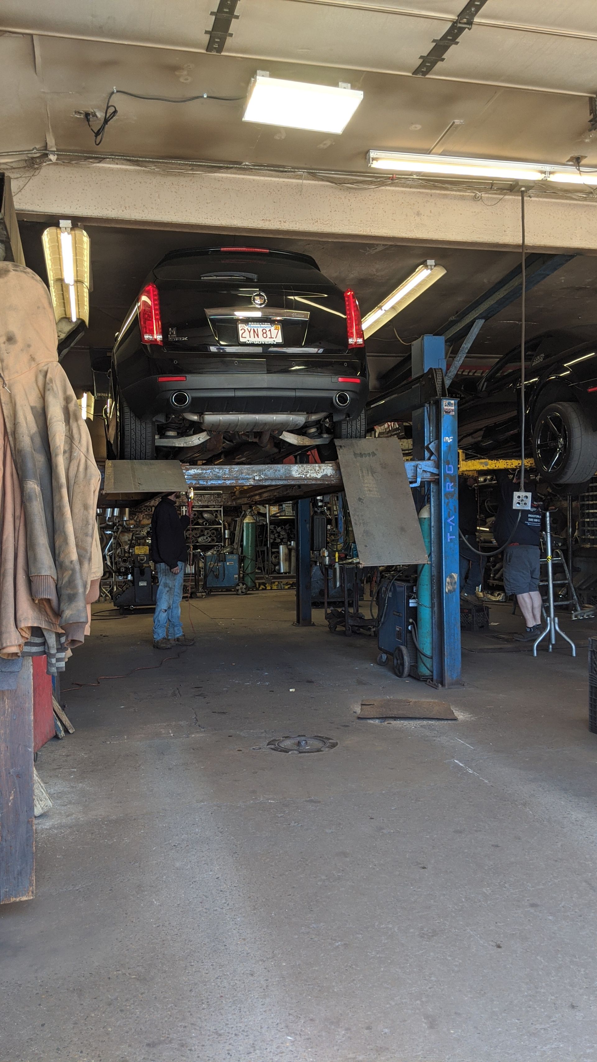 Car in a garage lift bay, raised on blue hydraulic posts with another vehicle in the background.