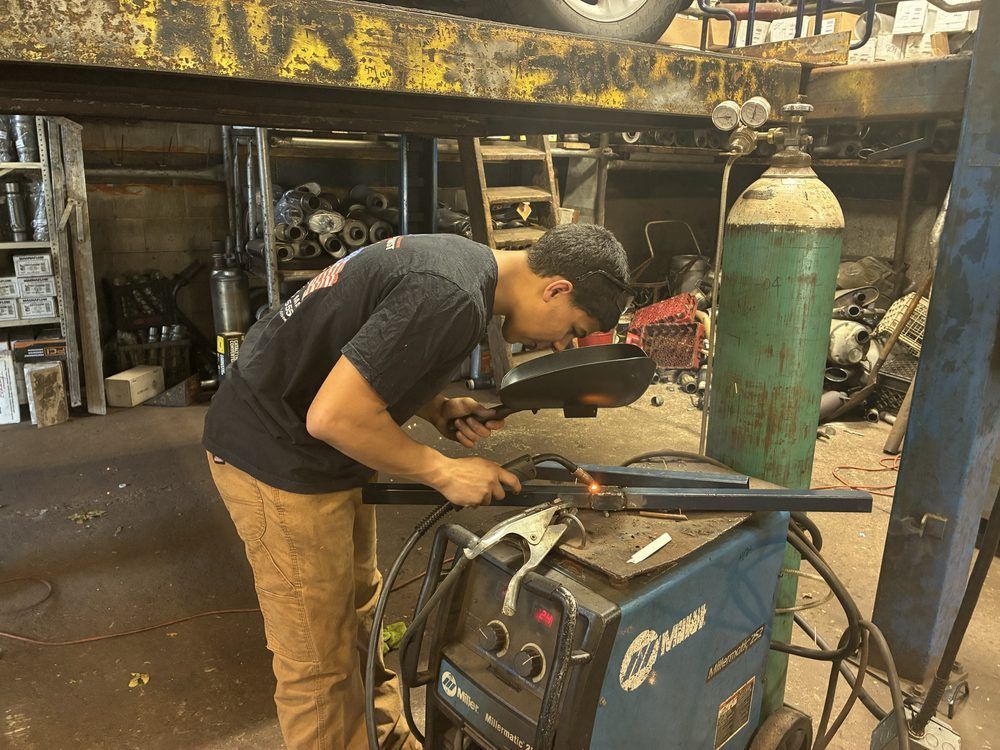 Person welding metal in a workshop beside a blue welding machine and green gas cylinder.