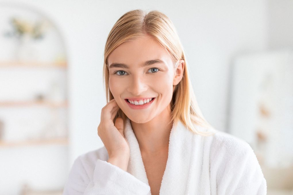 A woman in a white robe is smiling and touching her face.