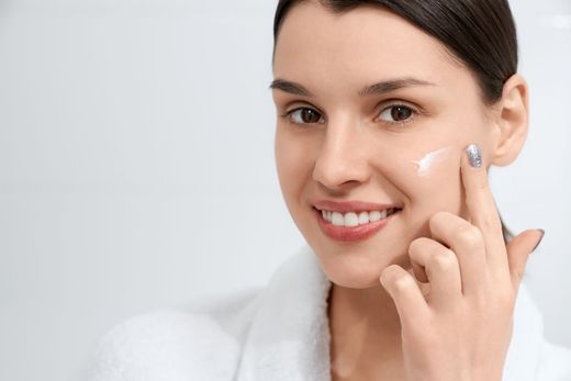 A woman is smiling while applying cream to her face.