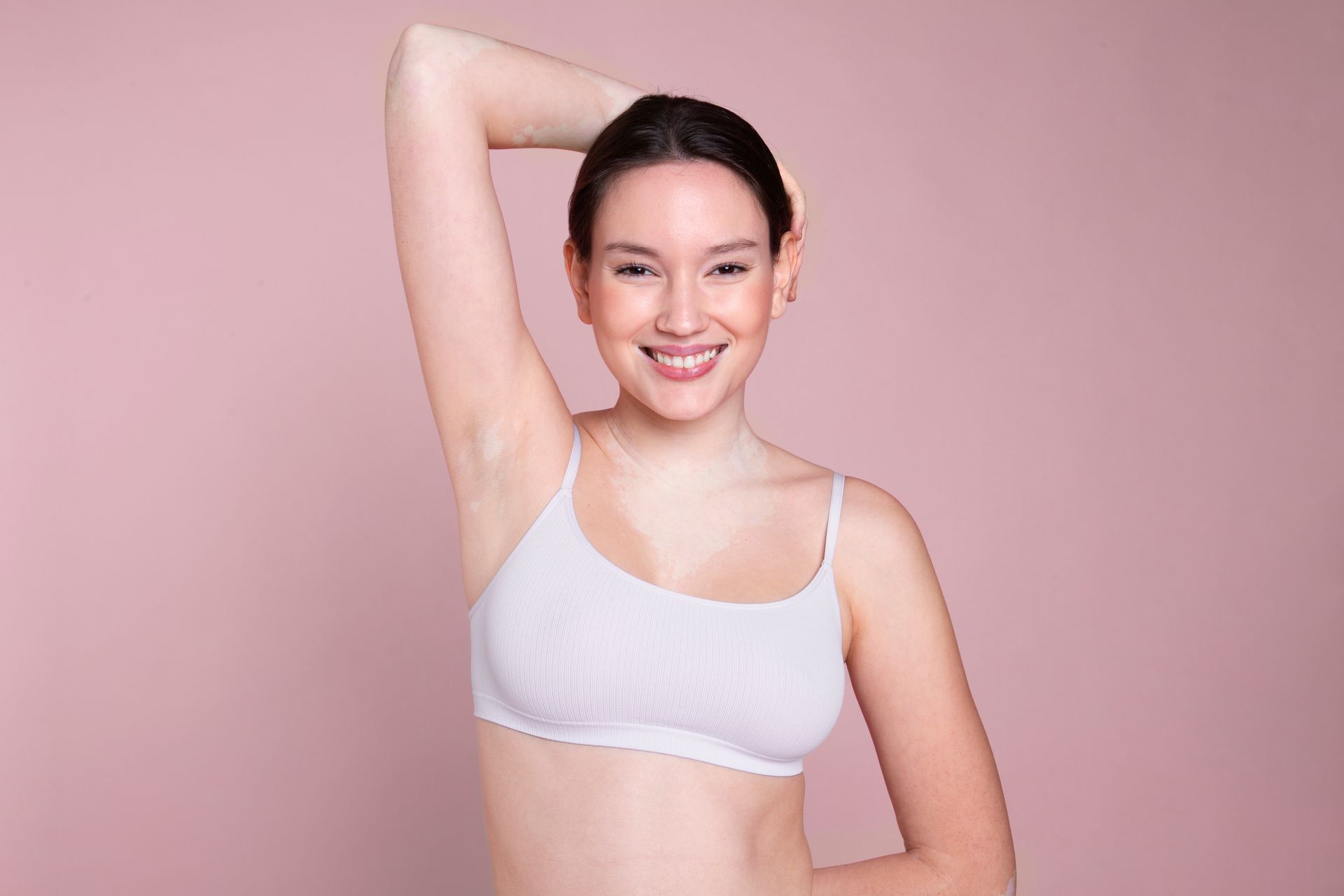 Woman in white bra smiles, arm raised to show underarm against a pink background.
