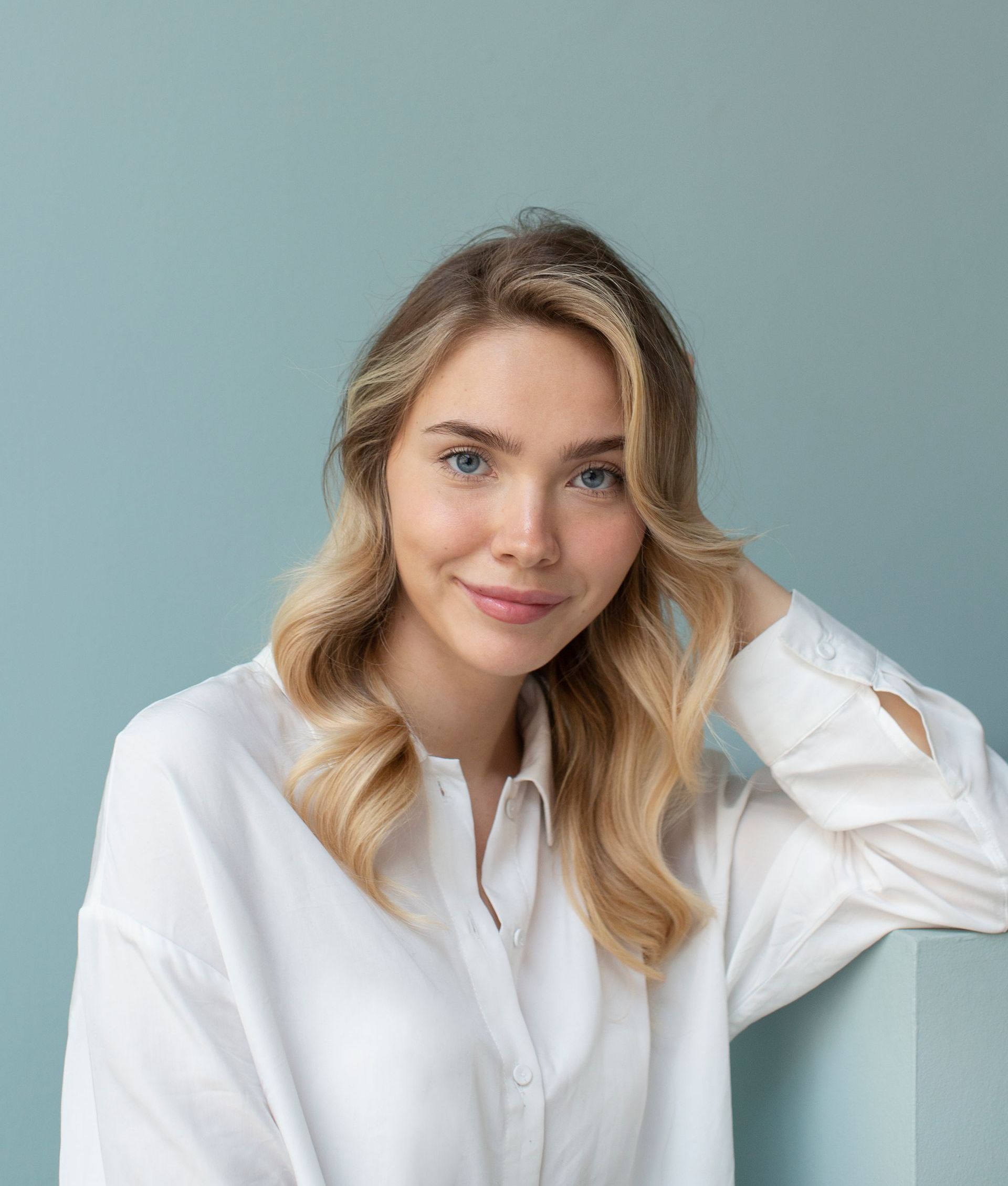 A woman in a white shirt is leaning on a wall and smiling.