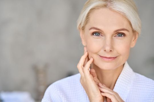 A woman in a white robe is smiling and touching her face.