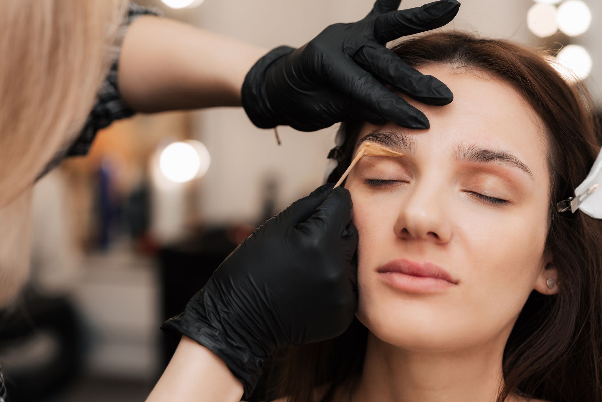 Person getting eyebrow treatment; black-gloved hands applying product. Close-up, eyes closed, beauty salon setting.