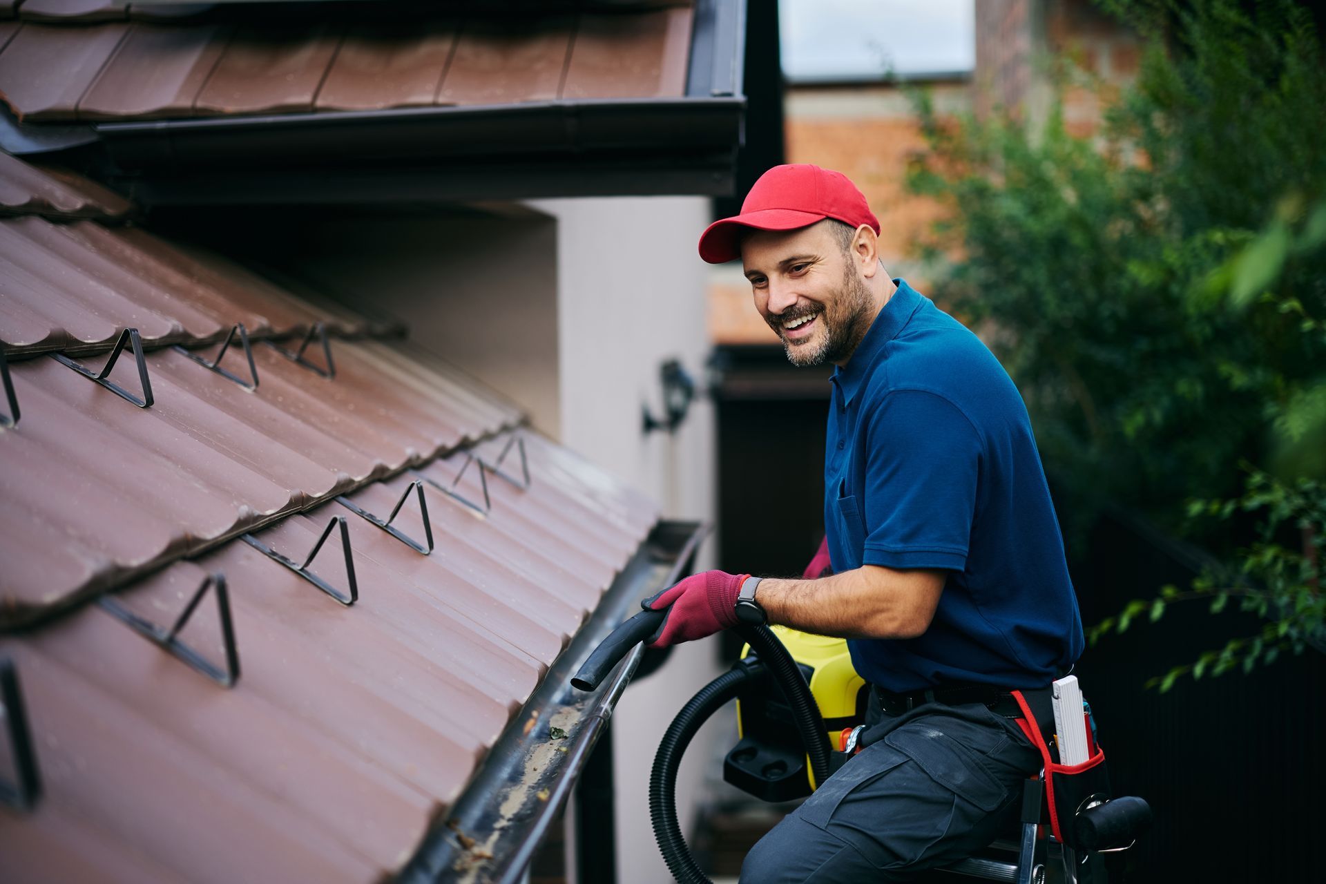 Man in red cap cleaning a brown roof with a pressure washer, smiling outdoors