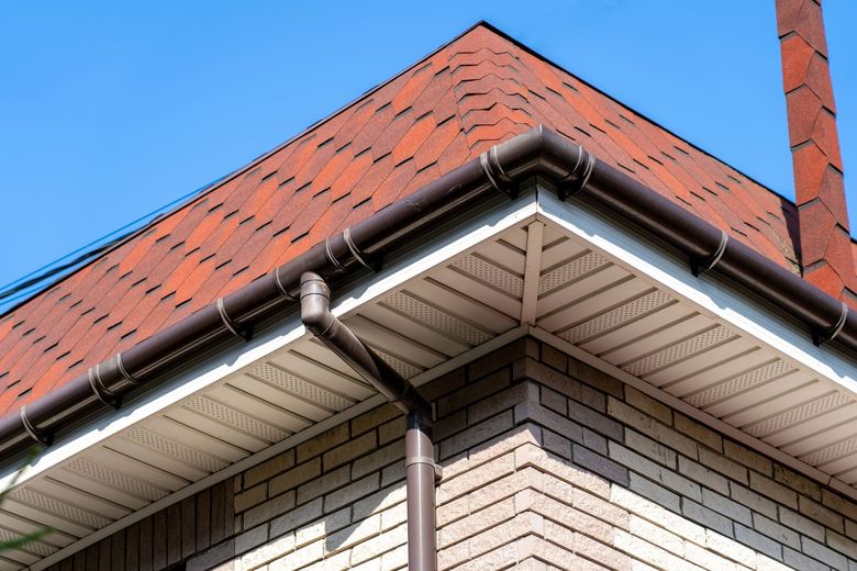 Corner of a brick house with a red tiled roof and visible gutter against a blue sky