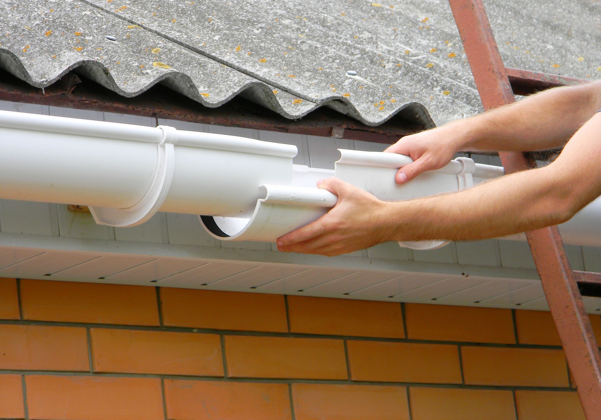 Hands installing a white gutter on a brick house under a corrugated roof.