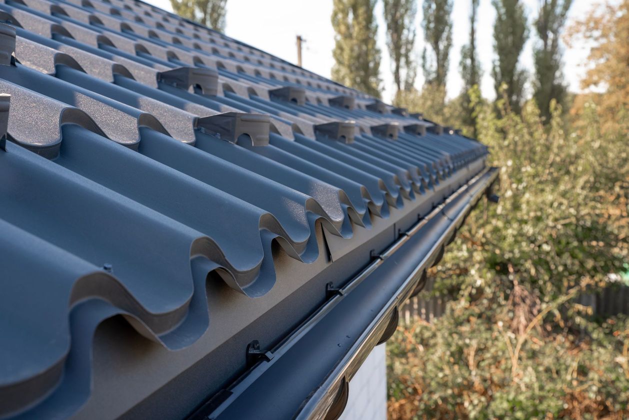 Close-up of a gray tiled roof with blue guttering beside trees and shrubs