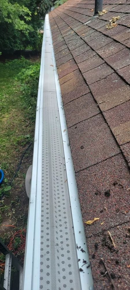 Roof edge with a white gutter and brown shingles, with leaves and debris along the roofline.