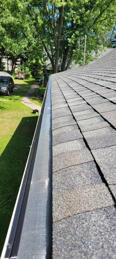 Roof shingles and gutter along a sloped roof beside a grassy yard and trees