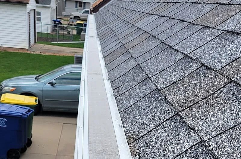Roof shingles and gutter on a house, with a driveway, parked car, and trash bins below.