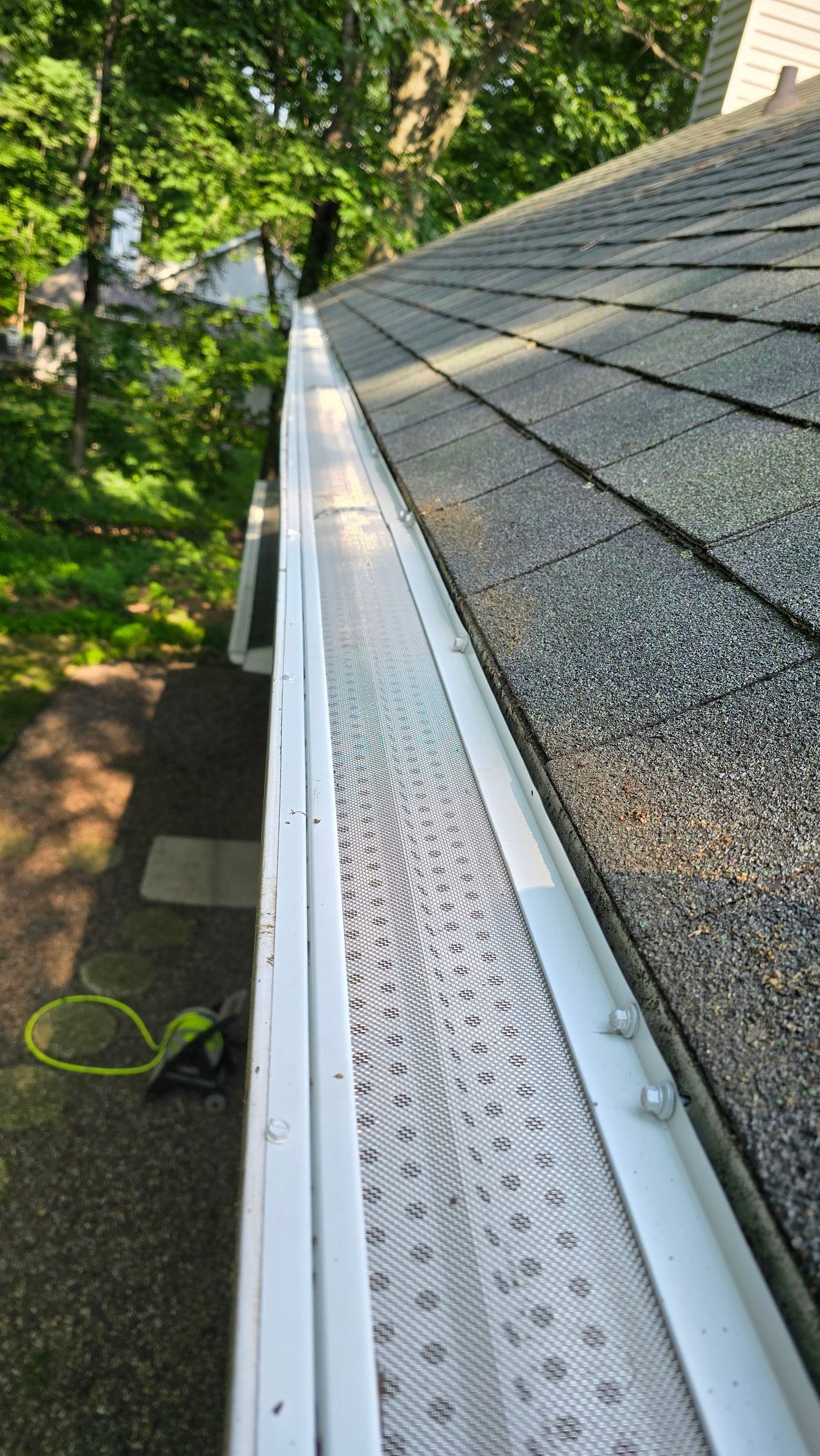Gutter guard along a shingled roof beside green trees and a yard below