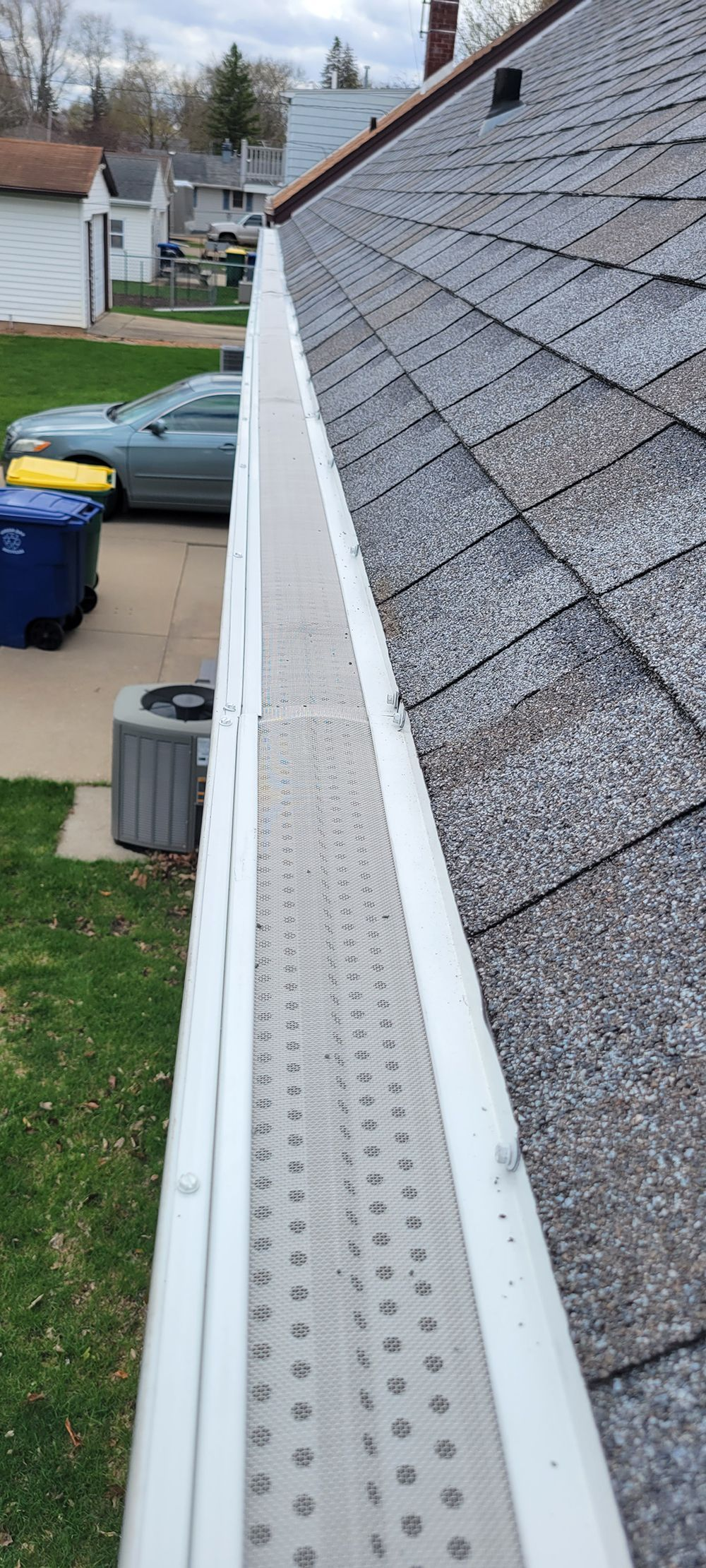 Close-up of a house roof and white gutter along an asphalt-shingled eave, with a driveway and trash bins below.