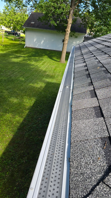 Roof edge with gray shingles and gutter beside a green lawn and a small tree under a bright sky