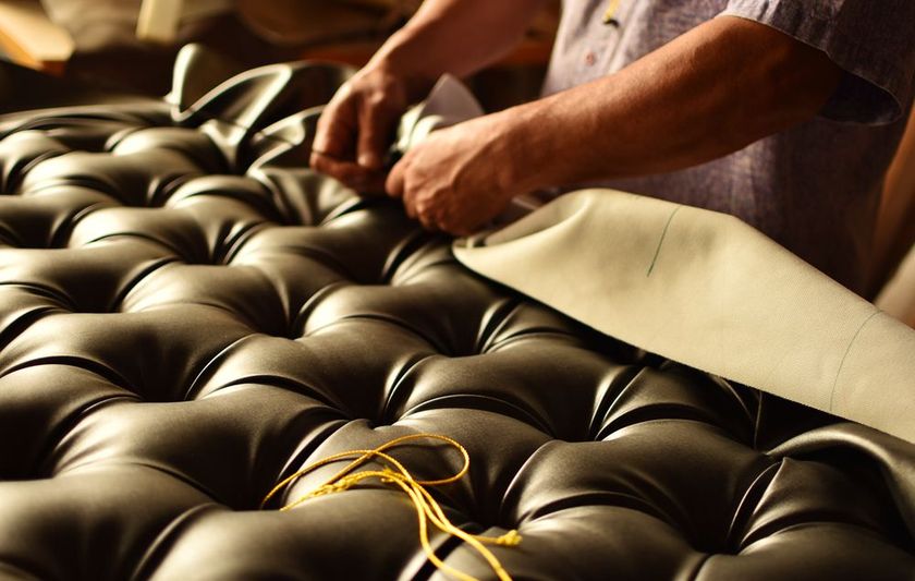 A craftsperson works on tufted black leather upholstery, carefully folding the fabric to create a diamond pattern.