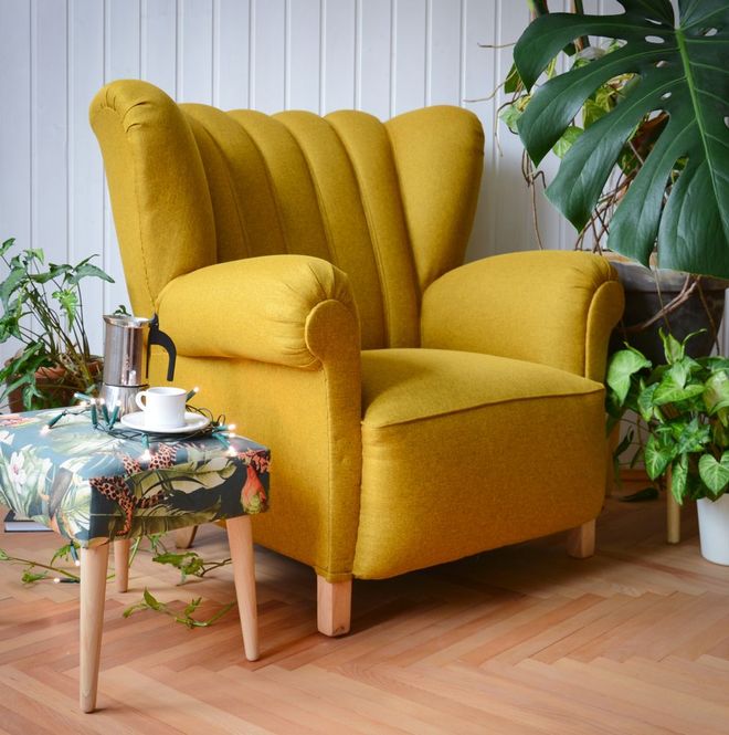 A mustard yellow channeled-back armchair with wooden legs sits on a wooden floor next to a small, floral-patterned stool.