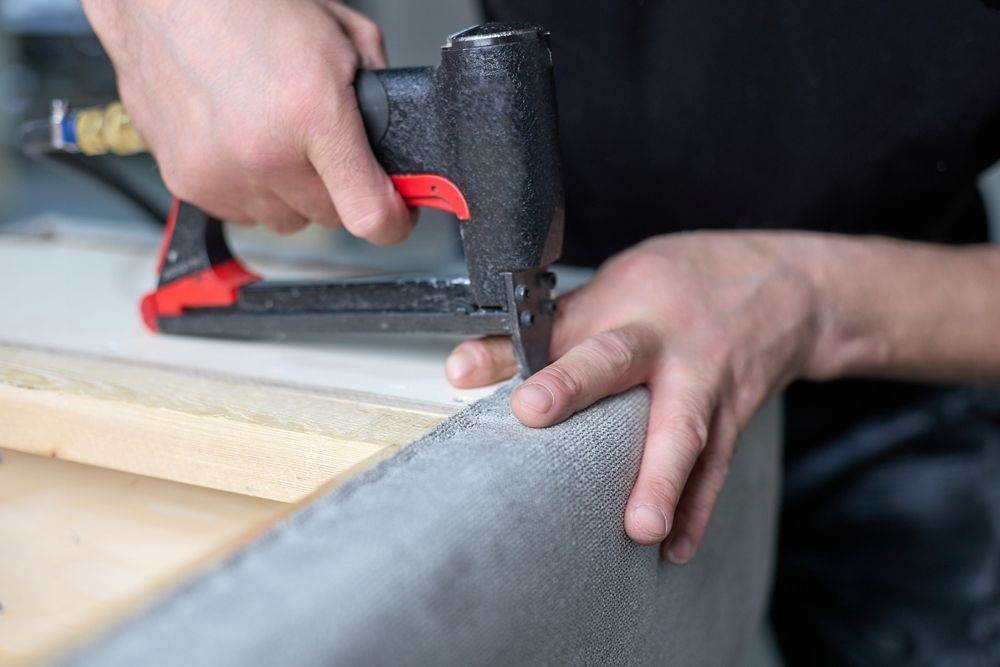 A person uses a pneumatic staple gun to attach gray fabric to a wooden furniture frame.