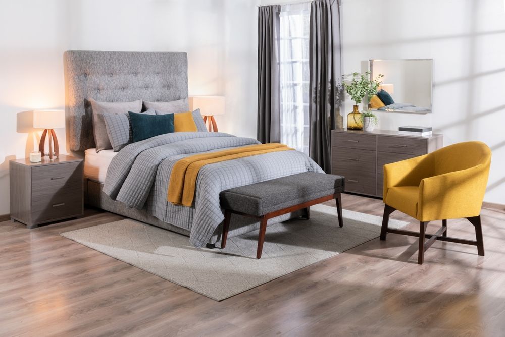 A modern bedroom featuring a gray tufted headboard bed, a yellow armchair, a wooden dresser, and matching nightstands.