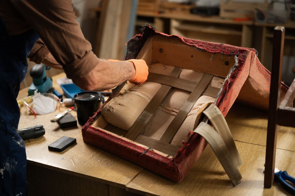 A person wearing an orange glove repairs a chair seat with webbing in a workshop.