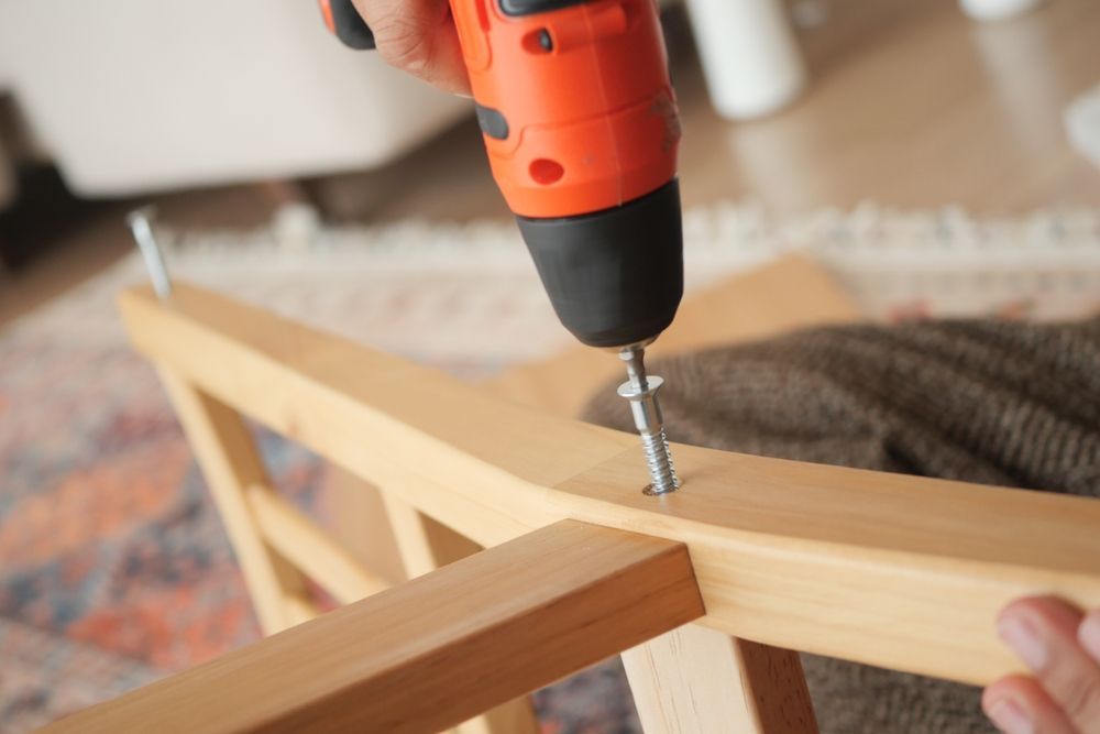 A person uses an orange electric drill to drive a screw into light-colored wooden furniture pieces.