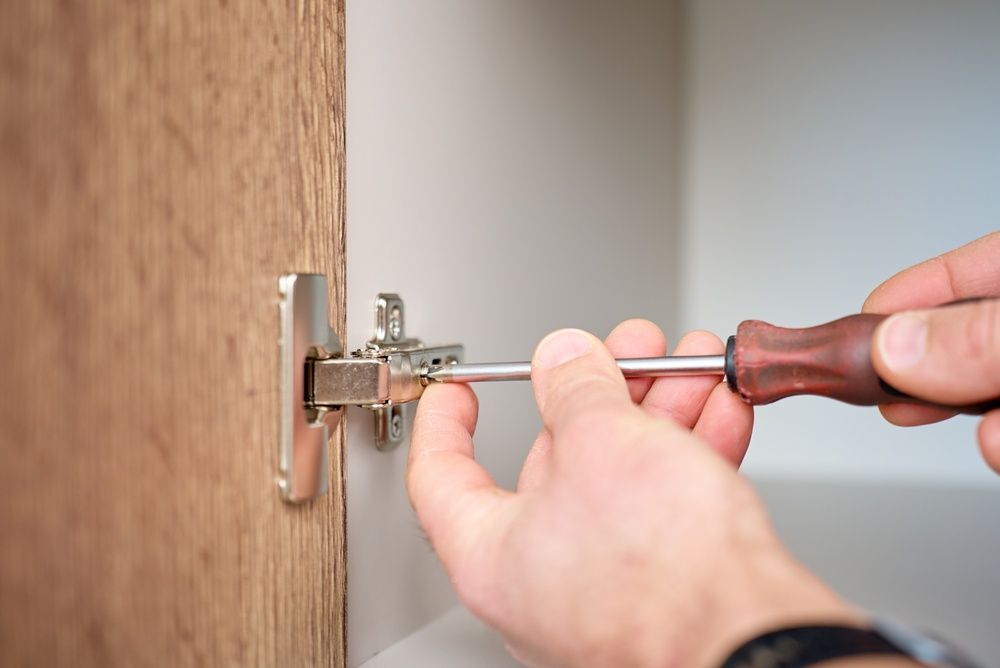 A person uses a brown-handled screwdriver to adjust a metal hinge attached to a wooden cabinet door.