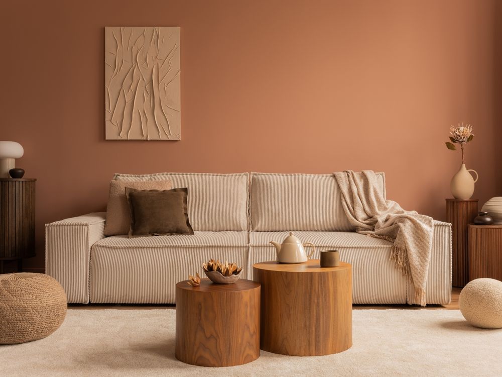 A beige striped sofa with a blanket, brown pillows, and wood coffee tables against a warm terracotta wall in a living room.