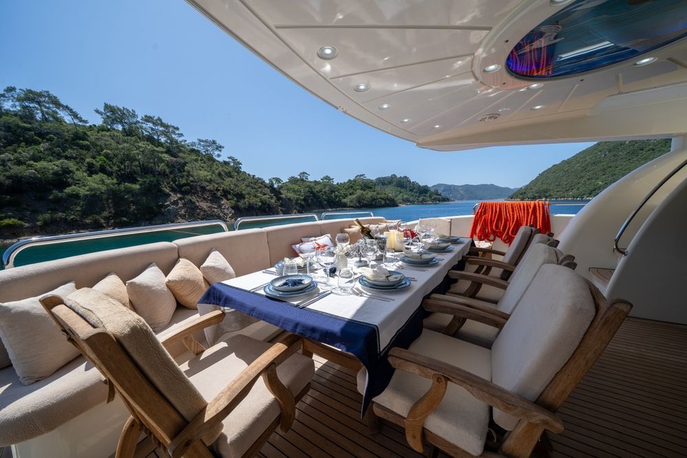 A dining table set for a meal on the outdoor deck of a yacht, overlooking a scenic bay with wooded hills.