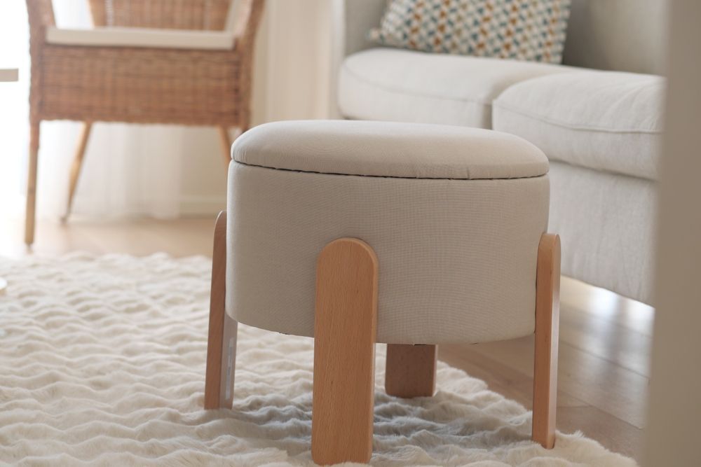 A cream-colored circular ottoman with light wooden legs stands on a textured white rug in a bright, modern living room.
