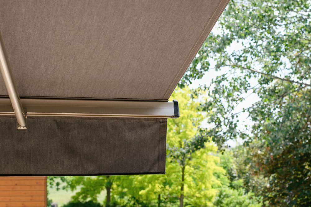 A tan fabric awning with a metal frame extends outward above a view of lush green trees on a sunny day.