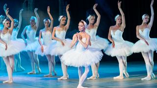 Ballet dancers in white tutus pose onstage under blue lighting, one centered in front with hands clasped.