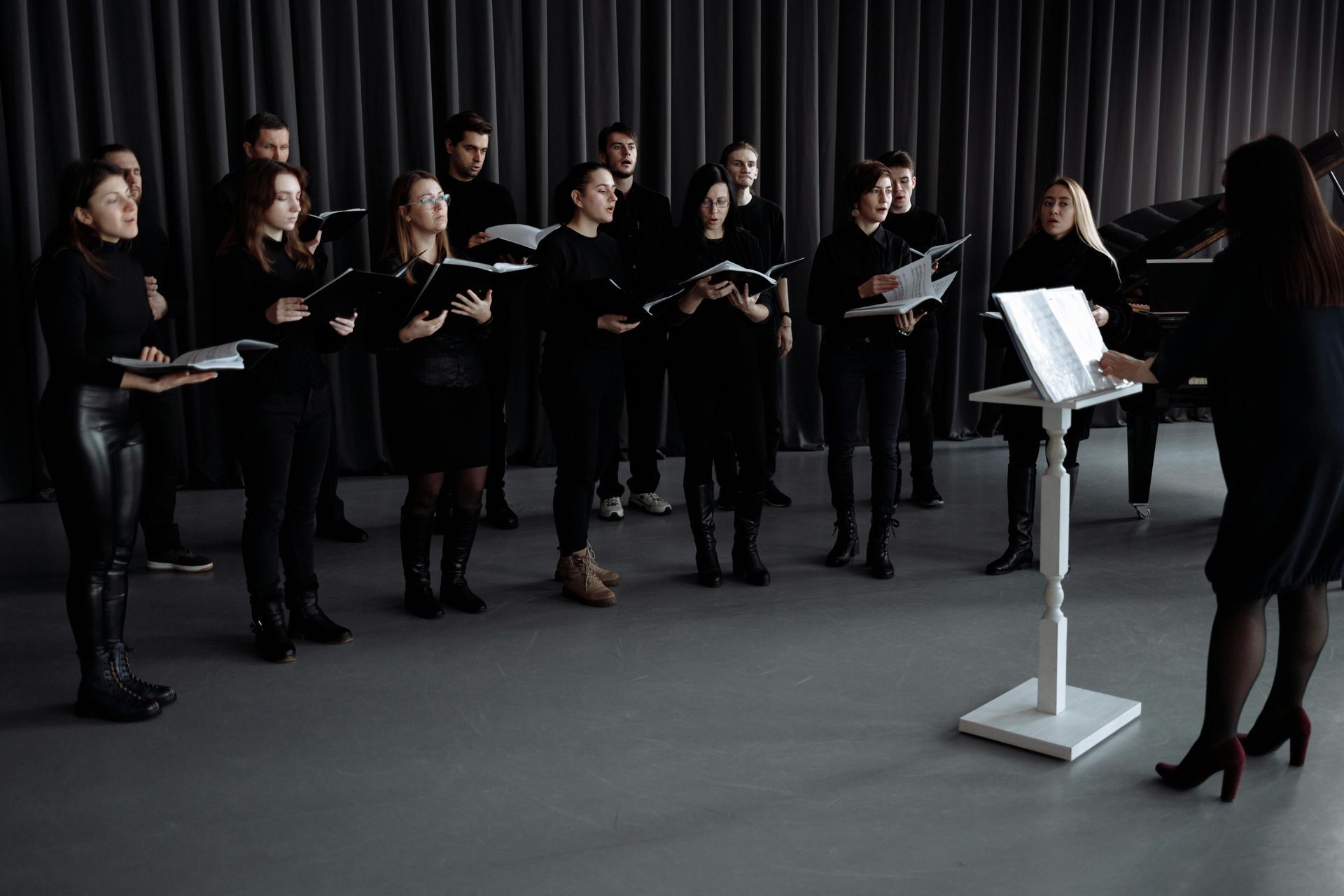 Choir rehearsing in a dark studio, reading from sheet music as a conductor leads them