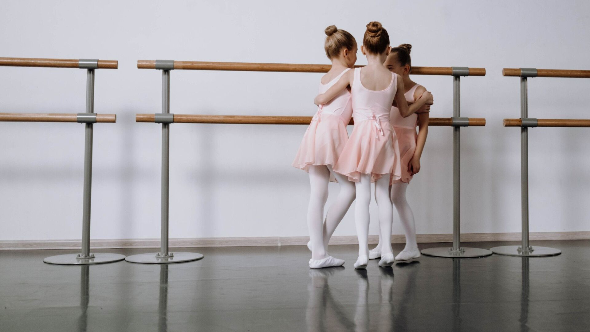Three ballet dancers in pink tutus and white tights stand at a barre in a studio.