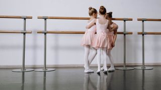 Three ballet dancers in pink tutus and white tights stand at a barre in a studio.