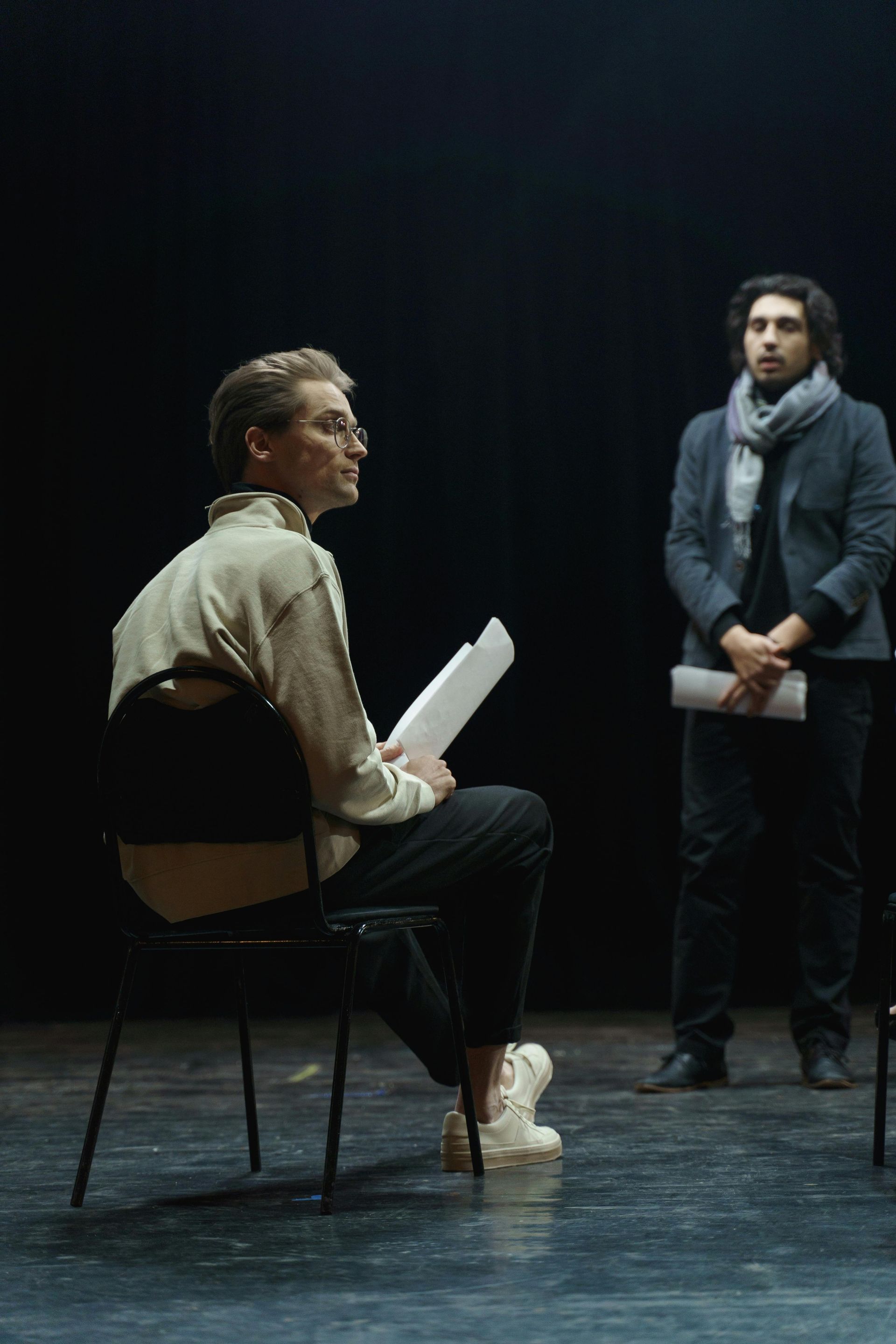 Two people on a dark stage, one seated holding papers, the other standing and speaking.